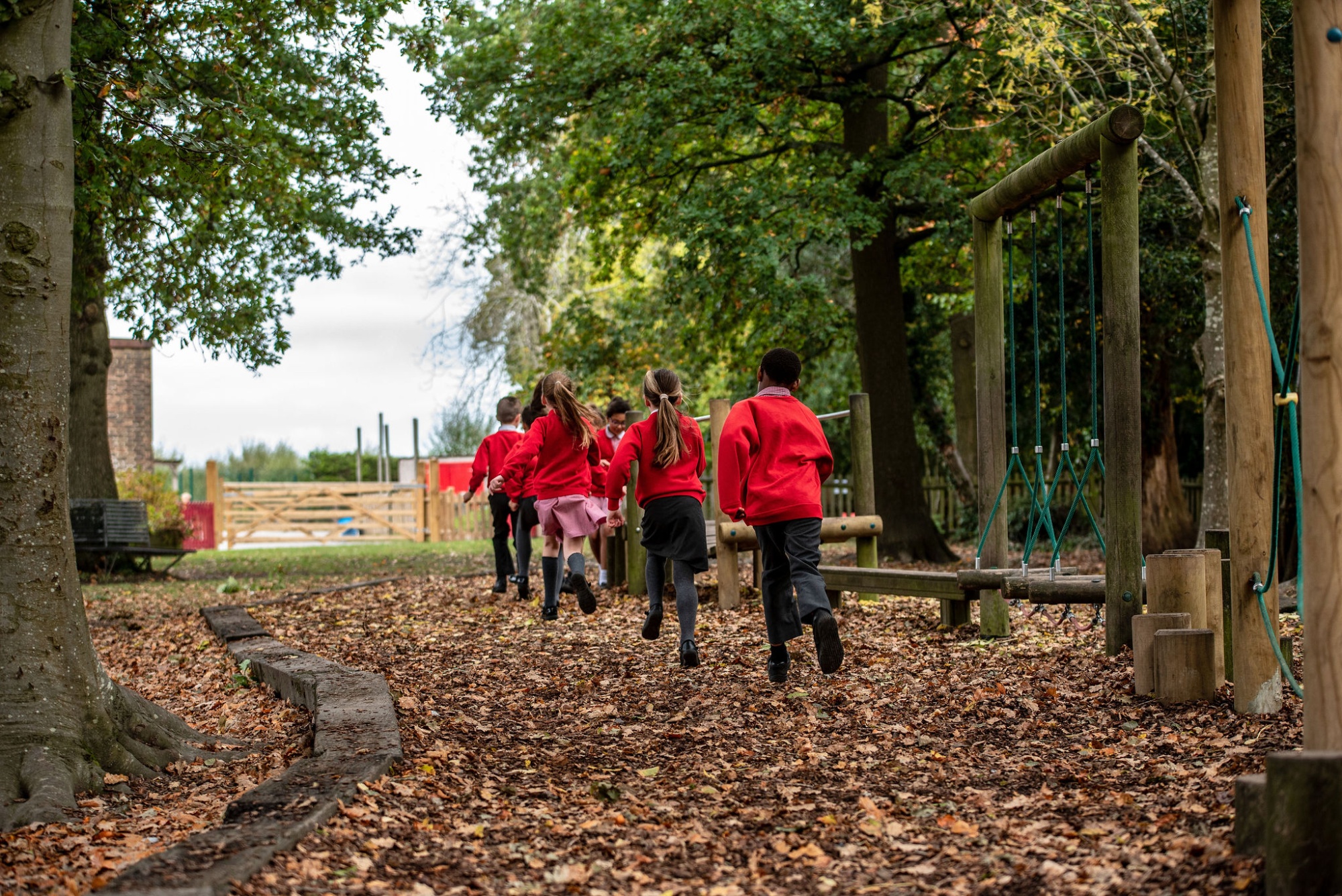 Five primary school children in red uniforms playing on outdoor adventure equipment in a wooded school playground